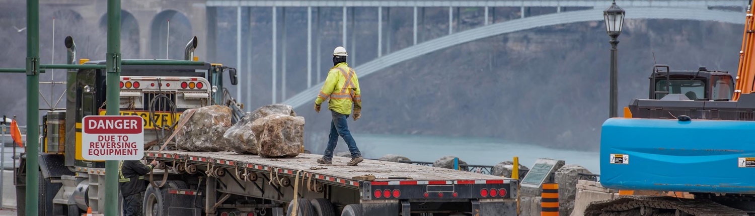 Construction worker on back of a semi truck with flatbed trailer loaded with large landscaping boulders